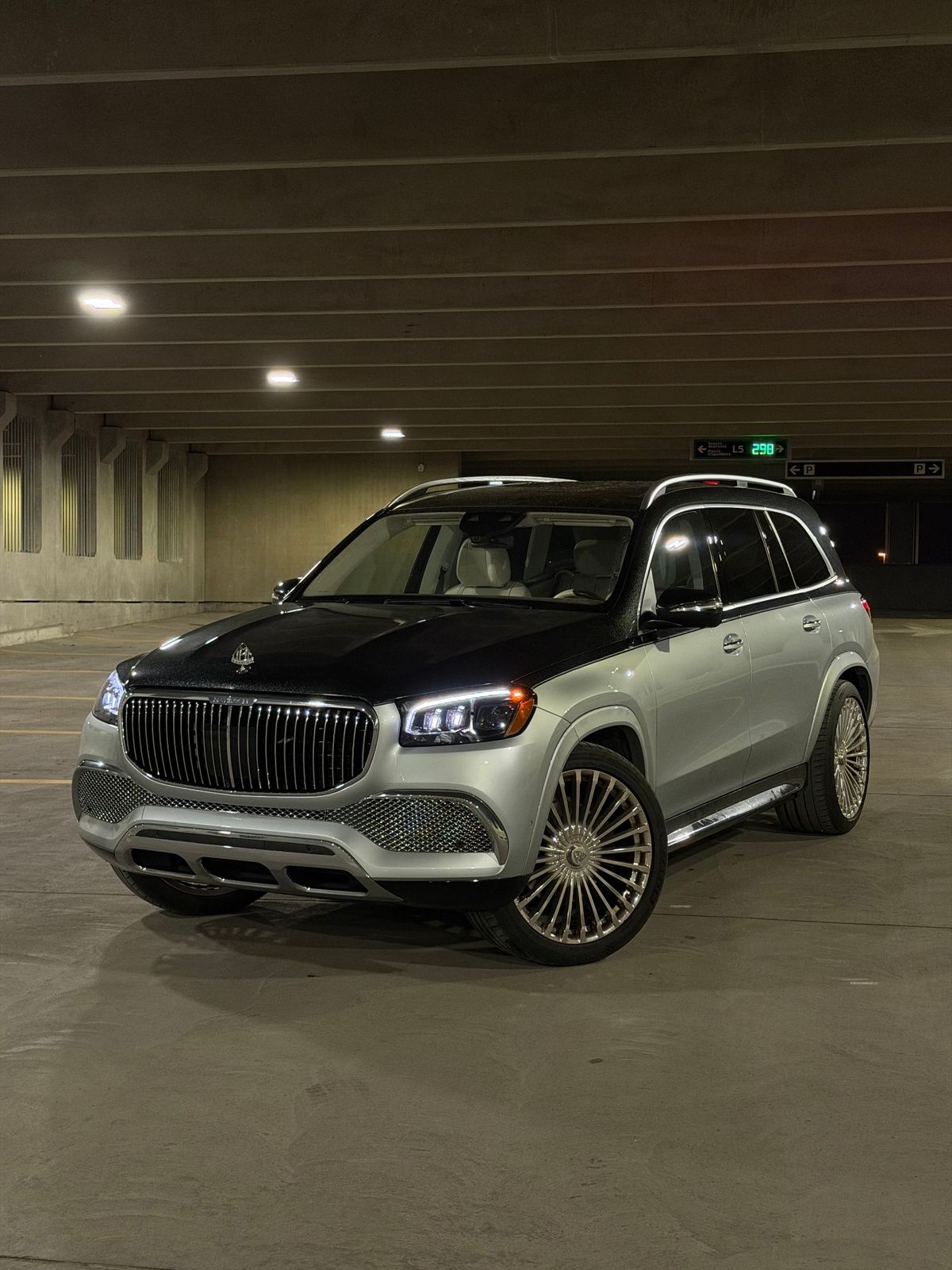 A silver and black Mercedes-Maybach GLS with its headlights on, inside a parking garage at night