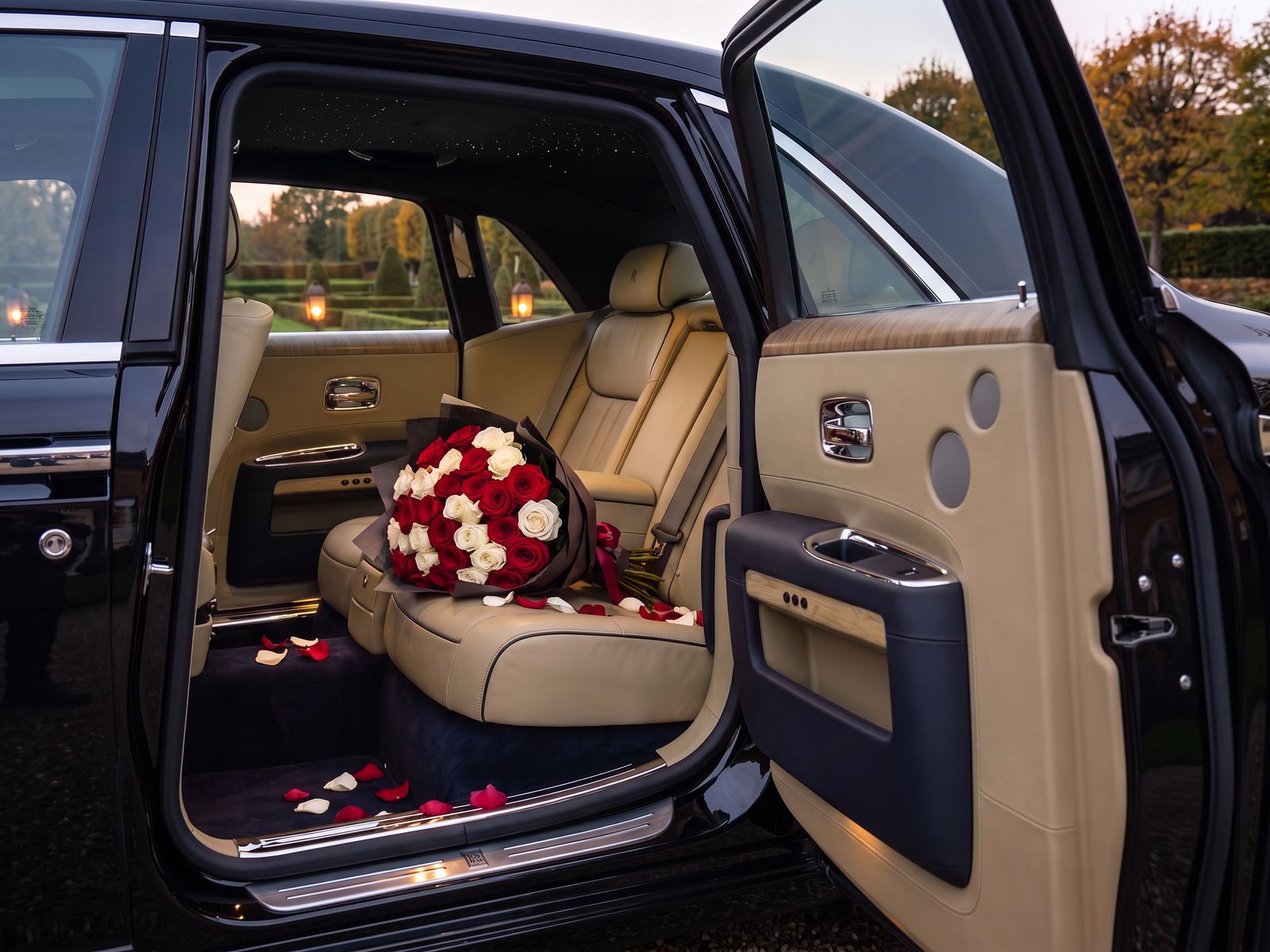 The open rear door of a black Rolls-Royce Ghost, with a bouquet of red and white roses on the tan leather rear seat and petals on the carpet